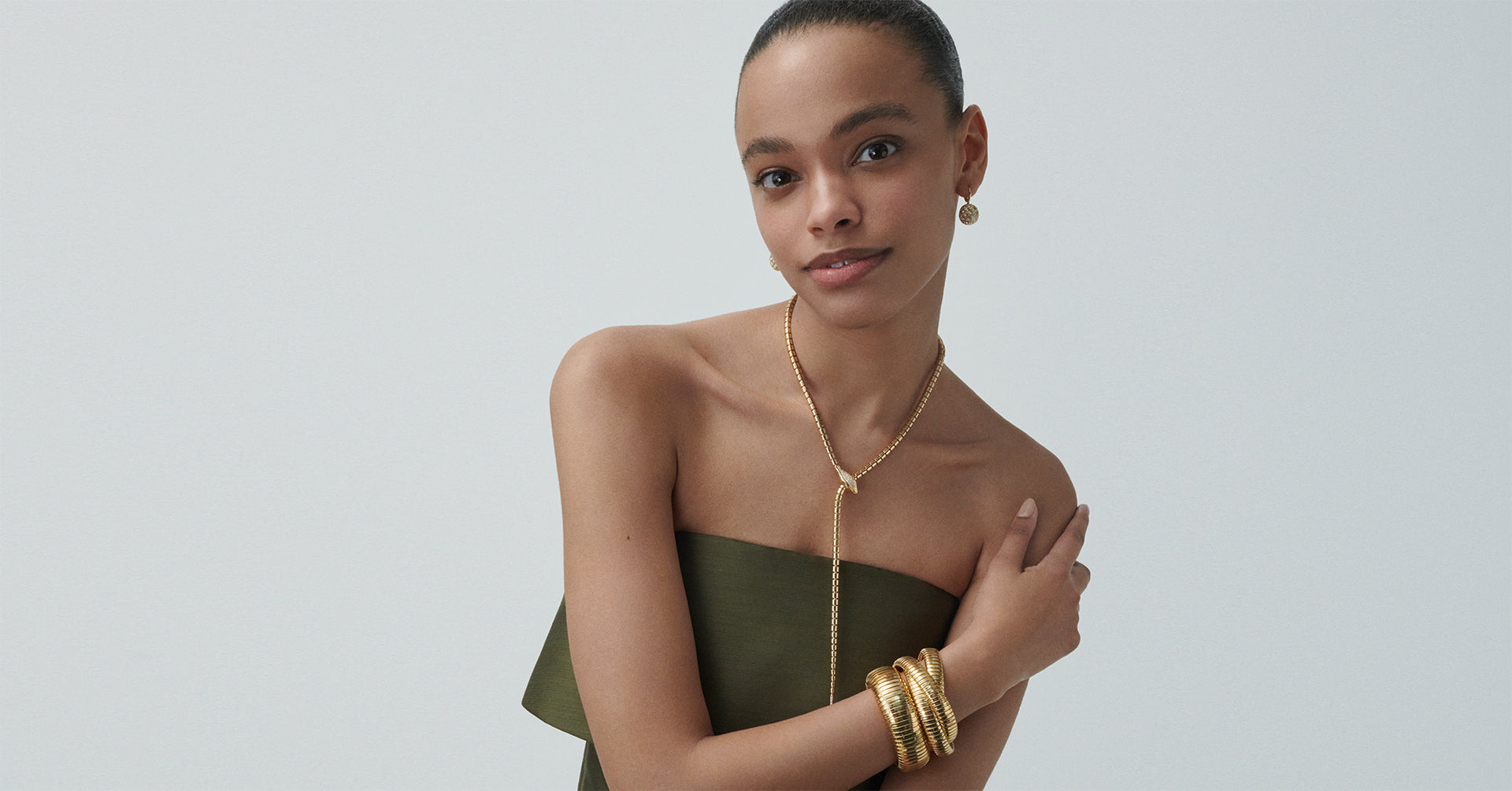 A model is wearing yellow gold honey comb earrings, wrap around necklace, and rolling bracelet and domed cuff stacked on her wrist.