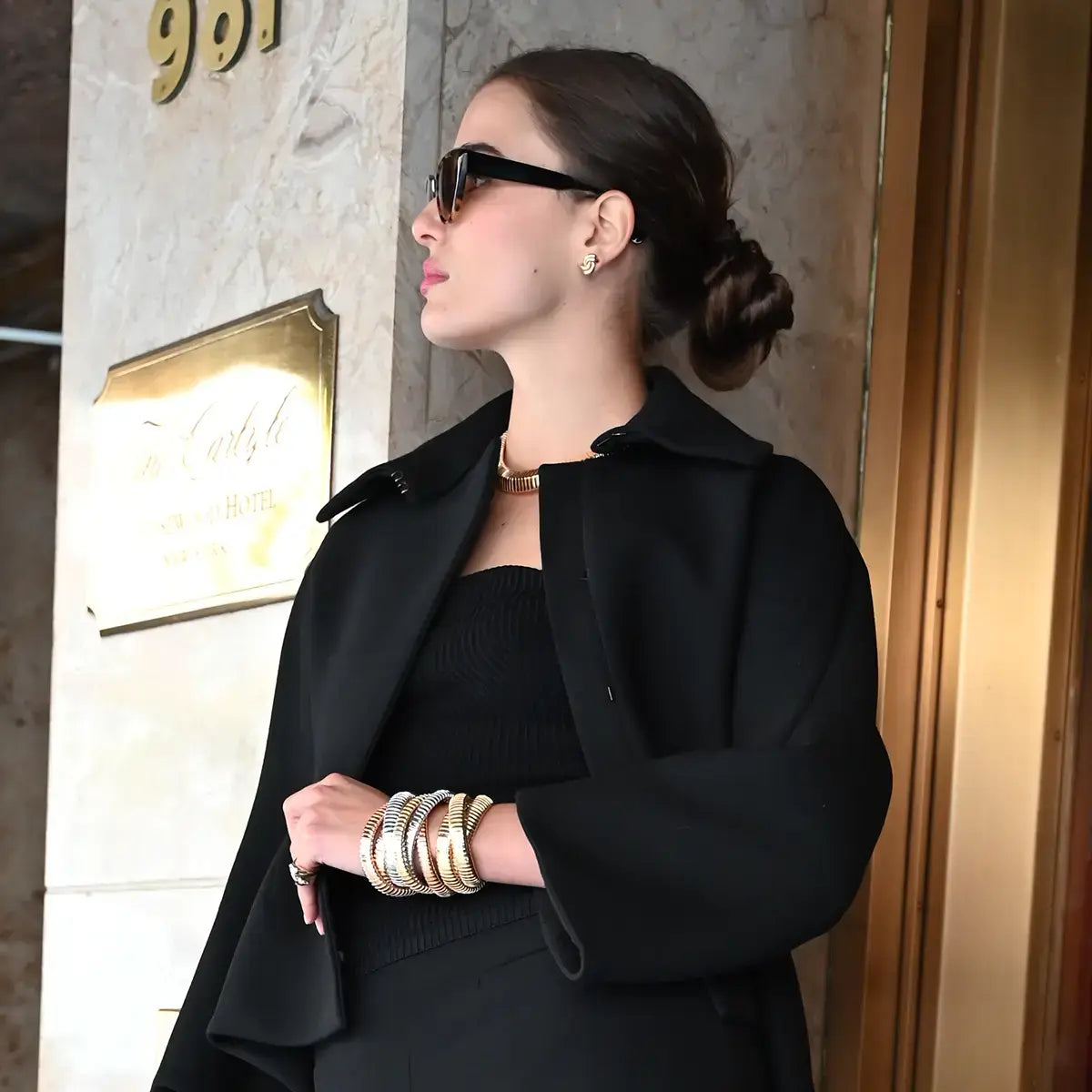 A model is wearing sunglasses, leaning against a wall and has on swirl earrings, rolling bracelets, a collana necklace and a serpente ring.