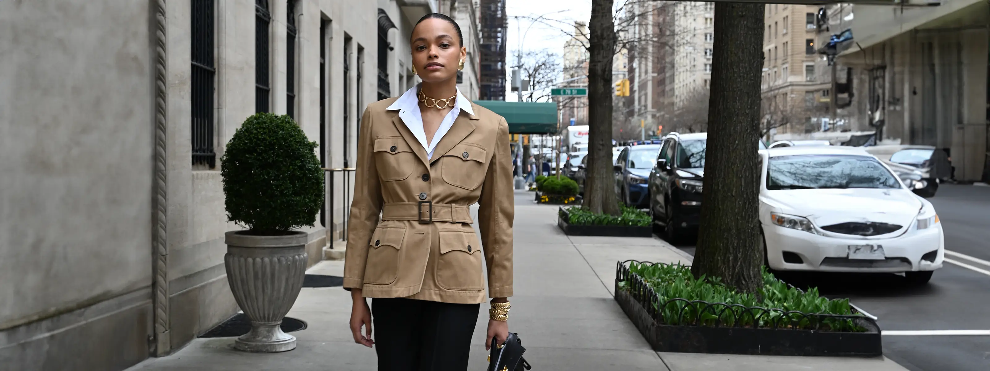 A model is walking down a city street and is wearing grooved anjou earrings in yellow gold, a yellow gold carre necklace and a stack of highly polished yellow gold wave link bracelets.