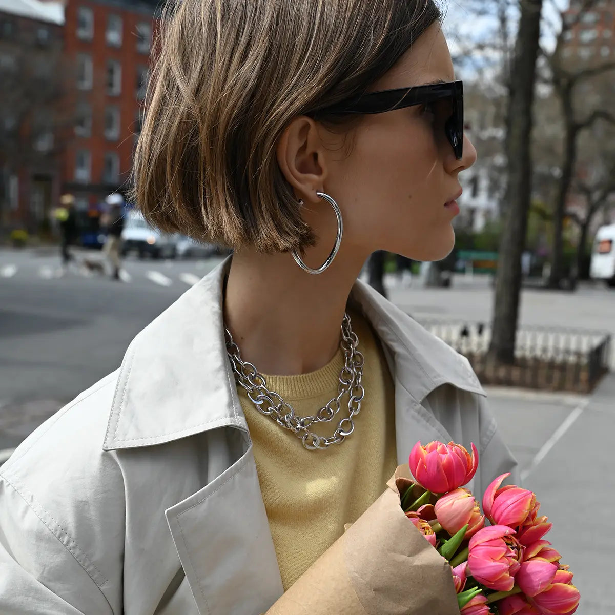 A model is wearing sunglasses, white gold curved oval hoop earrings, a white gold crescent link necklace, and holding tulips.