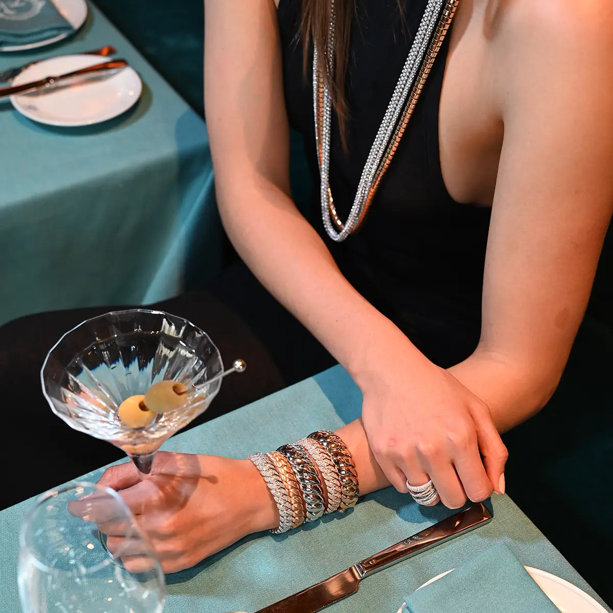 A model wearing rope and lunetta necklaces, a variety of wave link bracelets and a tall scribble ring with diamonds is seated at a table and is holding a martini.