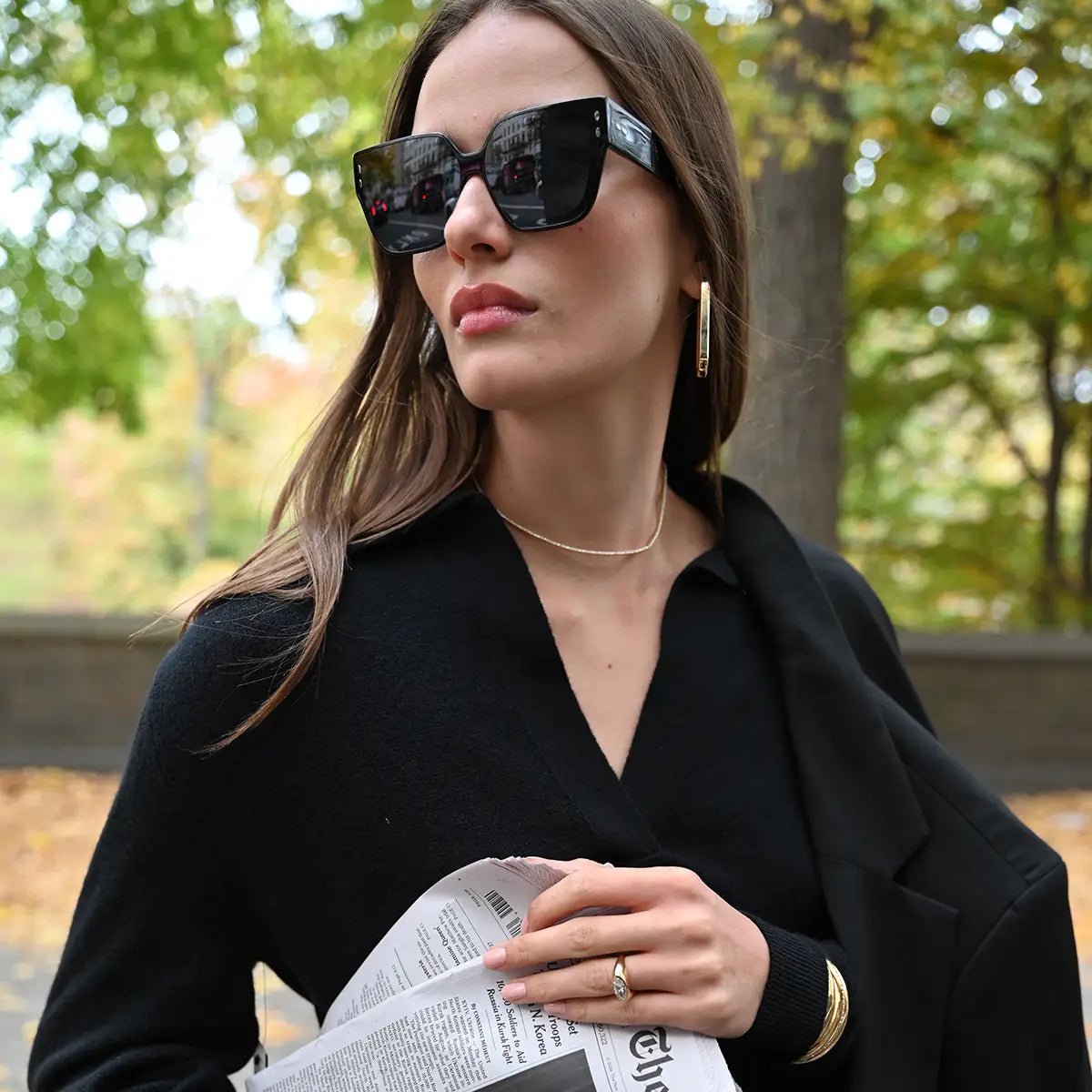 A model is wearing sunglasses and holding a newspaper. She is wearing a diamond accent scribble cuff, a marquise diamond ring, a lyra diamond necklace and wide paperclip hoops.