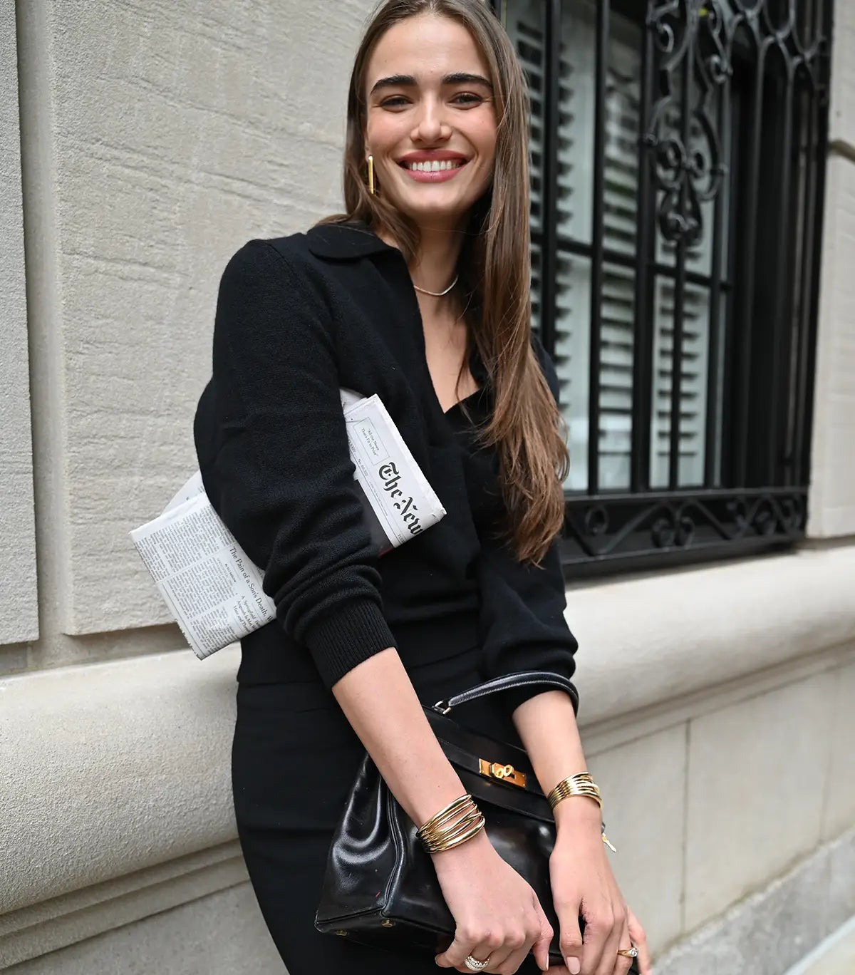 A model is smiling as she leans against a building facade with a copy of The New York Times tucked under her arm. She is wearing wide paperclip hoop earrings, a lyra necklace, and scribble cuffs on each wrist. She is also wearing french dome rings and a Carey ring.