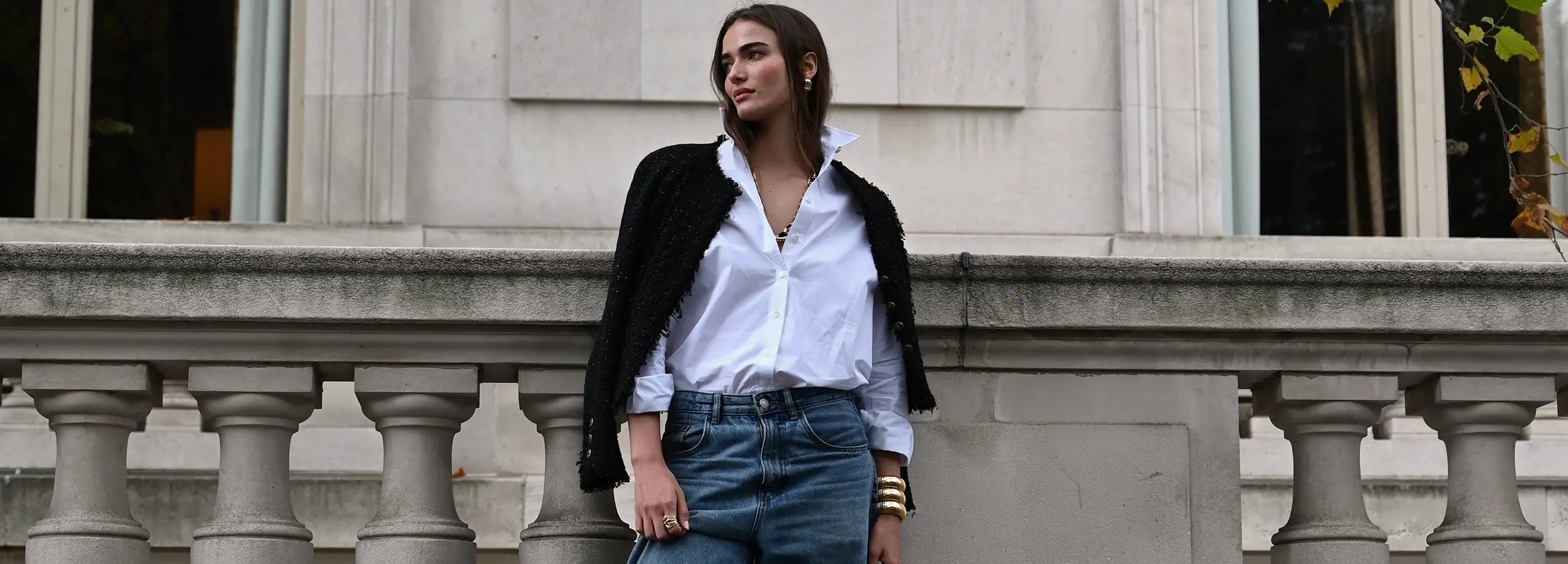 A model is wearing a black jacket and a white shirt, with a serpente ring, three yellow gold domed cuffs, and square clip on earrings. She is leaning against a stone wall.