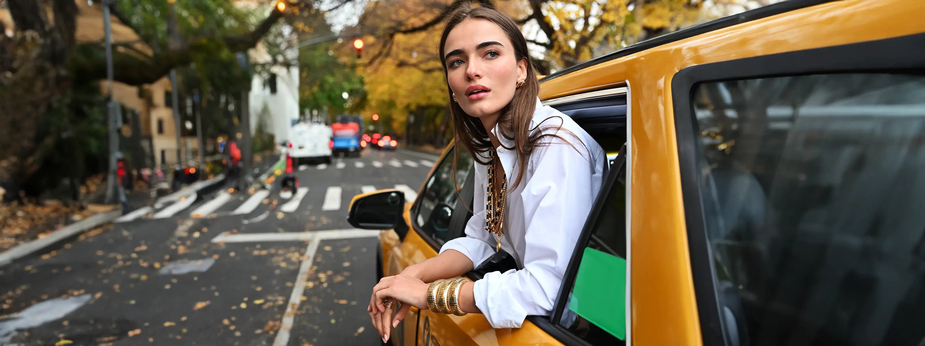 A model is riding in a yellow taxi and is wearing three yellow gold domed cuffs,  a black and white chain necklace, and square clip-on earrigns.