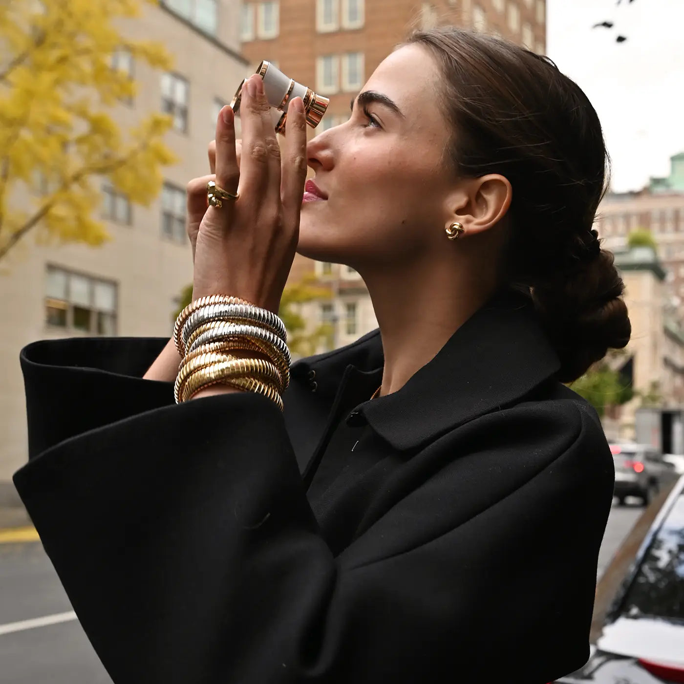 A model looks through opera glasses while wearing a yellow gold passionate panther ring, swirl earrings and rolling bracelets.