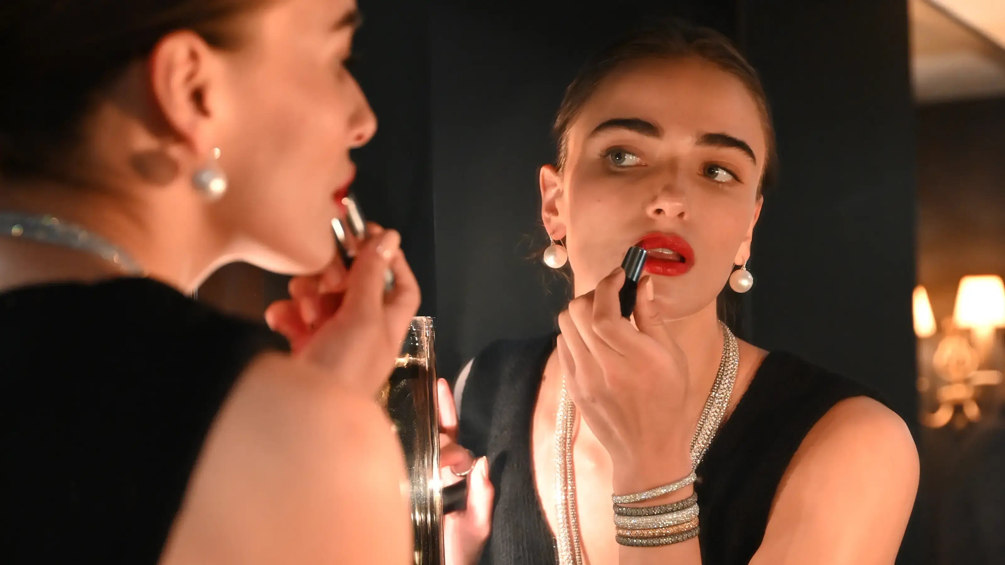 A model applying red lipstick into a mirror, wearing pearl earrings, diamond necklaces and diamond pave bracelets